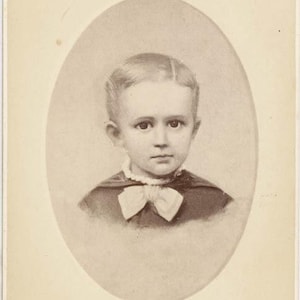May include: Sepia-toned portrait of a young child in an oval frame. The child has short, neatly combed hair, a white bow tie, and a dark-colored jacket. The image is a vintage photograph, likely from the late 19th or early 20th century.