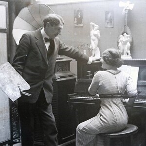 May include: A black and white photo of a man and a woman in a room. The man is holding a paint palette and is looking at the woman. The woman is sitting at a piano and is looking at the man. There are several sculptures on the wall behind them.