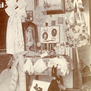 May include: An antique display featuring various vintage items. Includes framed portraits, a painting of boats, decorative linens, and silverware. The color palette is primarily sepia tones, creating a nostalgic feel. The items are arranged on a table and against a wall.