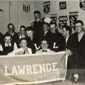 Może przedstawiać: Zdjecie w czerni i bieli przedstawiające grupę mężczyzn pozujących w pomieszczeniu. Z przodu trzymany jest transparent z napisem "LAWRENCE". Na ścianie widnieją proporczyki i znaki. Mężczyźni mają na sobie różne stroje.