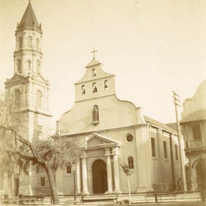 May include: A sepia photograph of a church, featuring a tall bell tower and a main building with a cross. The church has a classical facade with columns and a statue above the entrance. A tree is in the foreground.