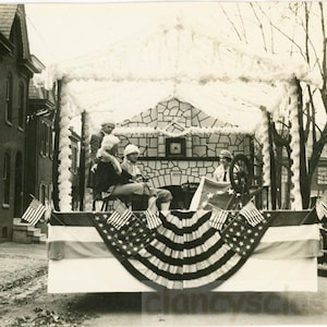 May include: A black and white photograph of a parade float decorated with white feathers and American flags. The float features a small stone building with a sign that reads "1776". People are sitting on the float, dressed in period clothing.