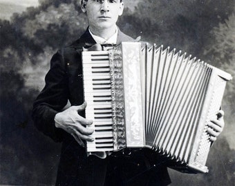 May include: A black and white photo of a man in a suit holding a large accordion. He is looking directly at the camera.