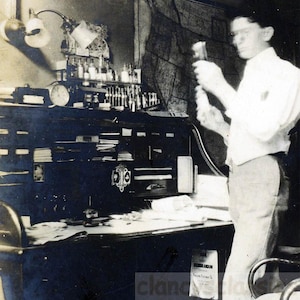 May include: Vintage black and white photograph of a man in a white shirt holding a glass vial in a laboratory setting. The lab features a desk with various bottles, a lamp, and a clock. A map hangs on the wall.