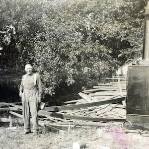 May include: Vintage black and white photograph of a sawmill operation. Three men are present, one standing with an axe, another operating a circular saw, and a third standing nearby. A steam engine with a tall smokestack is visible.