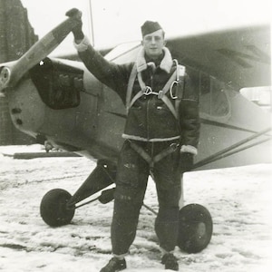May include: A vintage black and white photograph of a pilot standing next to a small airplane. The pilot is wearing a dark jacket with a fur collar, dark pants, and a cap. The plane has a propeller and a single wing.
