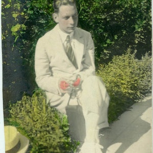 May include: A man in a white suit sits on a ledge with a leafy background. He is holding a red object in his hands.