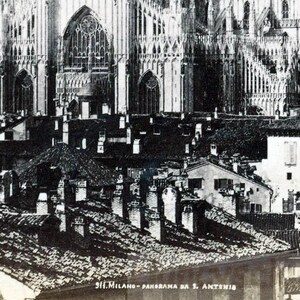 May include: Black and white vintage photograph of a cityscape featuring a large cathedral in the background. The foreground shows rooftops with chimneys. The image is labeled "911. Milano - Panorama da S. Antonio."