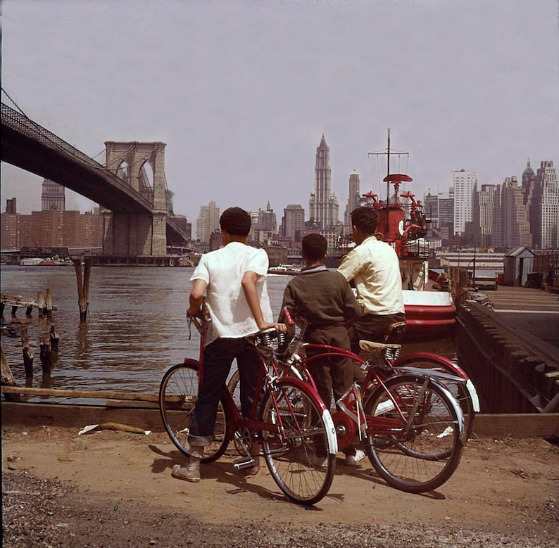 Puede incluir: Tres j&oacute;venes est&aacute;n de pie en un muelle frente al puente de Brooklyn, mirando hacia el agua. Cada uno lleva una bicicleta roja. El horizonte de la ciudad es visible en el fondo.