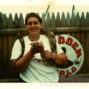 May include: A woman with long brown hair is holding a snake in her hands. She is wearing a white t-shirt with a red print that says "The Hidden World". The snake is brown and white and is wrapped around her arm. The woman is smiling and looking at the snake.