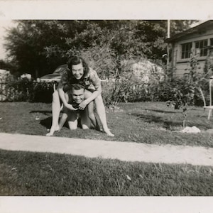 May include: A black and white photo of a couple playfully posing in a grassy yard. The woman is standing on the man's back, both smiling at the camera.