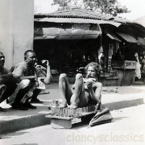 May include: A man sits on a bed of nails, with his legs crossed and his arms folded. He is wearing a white cloth around his waist. Two other men are standing behind him, watching. The scene is set in a street in India.