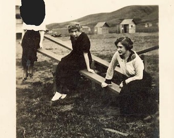 May include: Vintage black and white photograph featuring three people on a seesaw. The photo is set outdoors with a grassy foreground, a building, and a rural landscape in the background. The image has a classic, nostalgic feel.