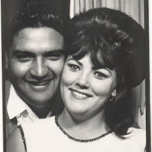 May include: Black and white vintage photograph of a smiling couple. The woman wears a white dress with a black trim. The man is wearing a collared shirt and a suit jacket. The photo is from clancysclassics.