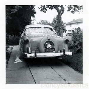 May include: A black and white photograph of a vintage car decorated with a white ribbon. The car has a number plate that reads "GA 84765".