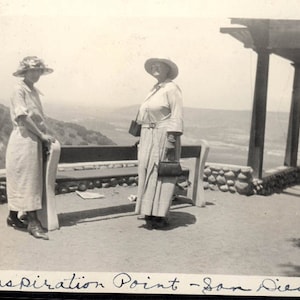 May include: Two women wearing hats stand on a wooden platform overlooking the ocean. The platform has a wooden bench and a stone wall. The text "Inspiration Point - San Diego - 1925" is written on the bottom of the photo.