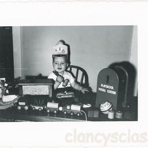 May include: A young child dressed as a mail carrier plays with a toy train set and a Playskool Postal Station. The child is wearing a black and white hat with a red stripe and a white shirt. The train set is made of wood and has a red, blue, and green engine. The Postal Station is made of red plastic and has a white label that says "Playskool Postal Station".