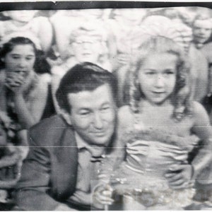 May include: A black and white photo of a group of people, including a man in a suit, a young girl in a dress, and a young boy in a hat. The man is smiling and looking at the camera. The girl is holding a doll and looking at the camera. The boy is looking at the camera.