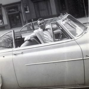 Może przedstawiać: Czarno-białe zdjęcie mężczyzny w garniturze siedzącego w kabriolecie. Samochód jest zaparkowany przed budynkiem z tabliczką z napisem "University Building".