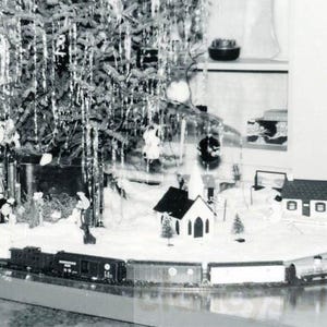 May include: Black and white photograph of a Christmas village scene. A decorated Christmas tree is in the background, with a miniature train set, houses, and figurines in the foreground. The scene is set on a table.