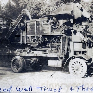 May include: Vintage black and white photograph of a Speed Well Truck and Thrasher. The truck has large wheels and is loaded with machinery. The image is taken outdoors with trees in the background.