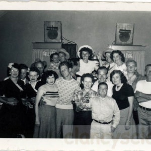 May include: A black and white photo of a group of people gathered together for a party. The group is standing in a room with a wooden floor and a few tables in the background. The people are all smiling and laughing, and they appear to be having a good time.