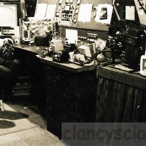 May include: A black and white photograph showing a workspace with electronic equipment, including a film projector, various radio devices, and a control panel with gauges. A person sits on a stool, working on a small electronic component at a desk.