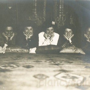 May include: A black and white vintage photograph of five young women sitting on a bed, smiling and looking at the camera. They are all wearing casual clothing and have their hands resting on their chins.