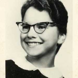 May include: A black and white portrait photograph featuring a person with short, wavy hair wearing dark-rimmed cat-eye glasses and a top with a rounded white collar. The text clancyclassics is visible in the bottom right corner of the image.