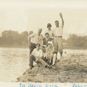 May include: A group of seven people stand and sit on a rocky outcrop by a river. The people are dressed in clothing from the 1920s. The text "Tri State Rocks Delaware, R.I." is written on the bottom of the image.