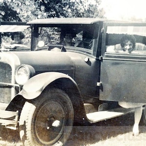 May include: A black vintage car with a wooden door and a woman sitting in the back seat. The car has a black grille and a spare tire on the back.