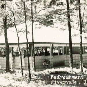 May include: A black and white photo of a refreshment stand in a wooded area. The stand has a roof and a brick wall. There are people standing in front of the stand. The text "Refreshment Stand Rivervale, Indiana" is printed below the image.