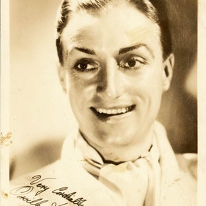 May include: A black and white photo of a man smiling with a white scarf around his neck. The photo is signed "Very Cordially, Orville Knapp."