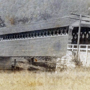 May include: A sepia-toned photograph of a covered bridge. The bridge has a wooden roof and sides, with decorative diamond-shaped cutouts along the eaves. A white railing runs along the side of the bridge, and a utility pole stands nearby.