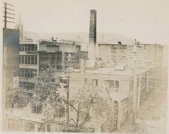 May include: A black and white photograph of a brick building with a tall smokestack. The building is surrounded by trees and power lines.