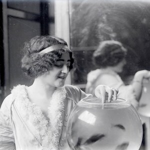 May include: A woman with dark hair and a headband is holding a glass bowl filled with water and fish. She is wearing a white, fluffy, fur-trimmed blouse. The bowl is on a table with a white tablecloth.