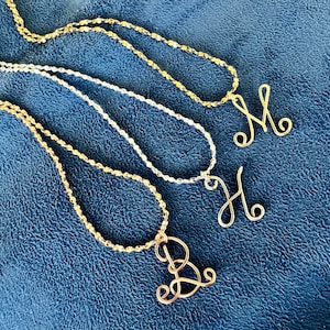 May include: Three gold necklaces with different initial charms. The charms are in the shape of the letters M, H, and D. The necklaces are on a blue fabric background.
