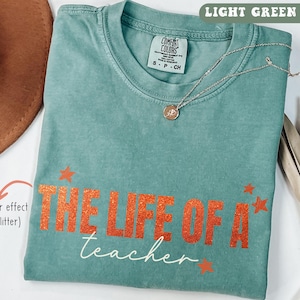 May include: Light green t-shirt with the text "THE LIFE OF A teacher" in orange glitter effect. The shirt has a crew neck and short sleeves. A silver necklace with a pendant and a brown hat are also visible.