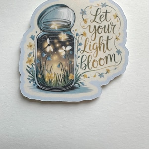 May include: A light blue sticker featuring a jar filled with glowing lights and flowers. The jar is labeled with the words "Let your Light Bloom" in a cursive font, surrounded by floral accents. The sticker has a light blue border.