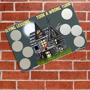 May include: A gray and black illustrated game card with a haunted house, a witch's legs, and the text "Flying Lessons Took A Wrong Turn". The card has five blank circles for game play and the text "Save $15".