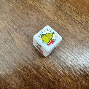 May include: A white six-sided die featuring a graphic of a green character with a grumpy expression surrounded by colorful Christmas lights on the top face, and a small illustration of a character in a red outfit on the side face.