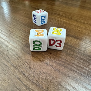 May include: Three white six-sided dice featuring colorful printed labels. One die shows D8 in blue and D12 in blue, another shows D4 in orange and D10 in green, and the third shows D6 in yellow and D3 in red. The dice are square with rounded edges.
