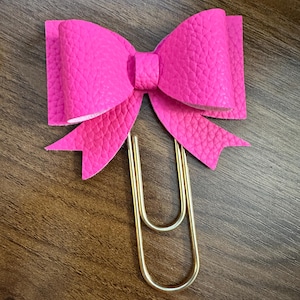 Op de afbeelding: Een felroze strikvormige paperclip. De strik is gemaakt van een textuurrijk materiaal en is bevestigd aan een goudkleurige metalen clip. De clip heeft een standaard formaat en vorm. De strik is gecentreerd op de clip.