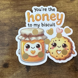 May include: A sticker featuring a jar of honey and a biscuit, both with cute faces. The text "You're the honey to my biscuit" is at the top. The jar is orange and the biscuit is yellow.