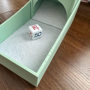 May include: A mint green dice tower with a felt-lined interior and an arched opening. A white die with red and blue numbers rests inside the tower. The numbers on the die are 25 and 50. The tower sits on a wooden surface.
