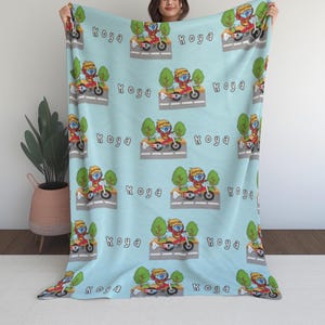 May include: Pale blue blanket with a repeating pattern of a cartoon character riding a motorcycle, accompanied by the word "Koya". The design includes trees and a road, creating a playful scene. The blanket is soft and inviting, perfect for a child's room.