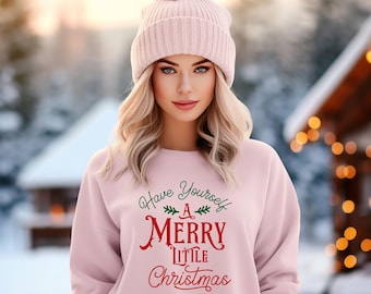 May include: A light pink sweatshirt with the text "Have Yourself A Merry Little Christmas" in green and red. The person is wearing a matching pink knit beanie with a pom-pom. The background is a snowy winter scene.