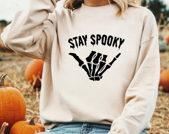 Może przedstawiać: Beżowa bluza z napisem "STAY SPOOKY" nad czarnym gestem dłoni szkieletu. Bluza ma neutralny kolor, idealny na jesień i Halloween. W tle widoczne są dynie.