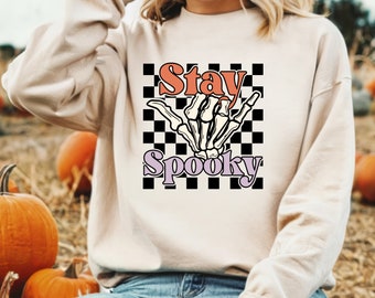 Może przedstawiać: Beżowa bluza z nadrukiem w stylu retro na Halloween. Grafika przedstawia szkielet dłoni z napisem "Stay Spooky" w kolorze pomarańczowym i fioletowym na czarno-białym tle w szachownicę.