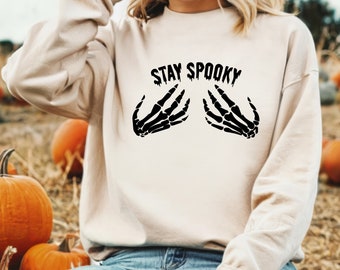 Może przedstawiać: Beżowa bluza z napisem "STAY SPOOKY" nad grafiką dwóch czarnych szkieletowych dłoni. Bluza z długim rękawem i okrągłym dekoltem jest idealna na Halloween lub na straszny sezon.